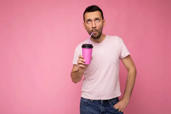 수염을 기르고 핑크 색 배낭 벽에 고립된 흰색 티셔츠를 입고 종이컵을 들고 마시고 즐기고 있는 잘생긴 유쾌 한 젊은 갈색 머리 남자 — 스톡 사진
