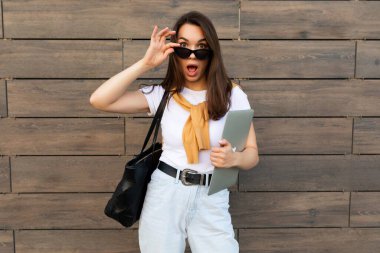 Büyüleyici, güzel, gülümseyen, çekici, esmer kız elinde bilgisayar bilgisayarı, elinde siyah güneş gözlüğü, beyaz tişörtlü, sarı kazaklı ve sokakta açık mavi kotlu kameraya bakıyor.