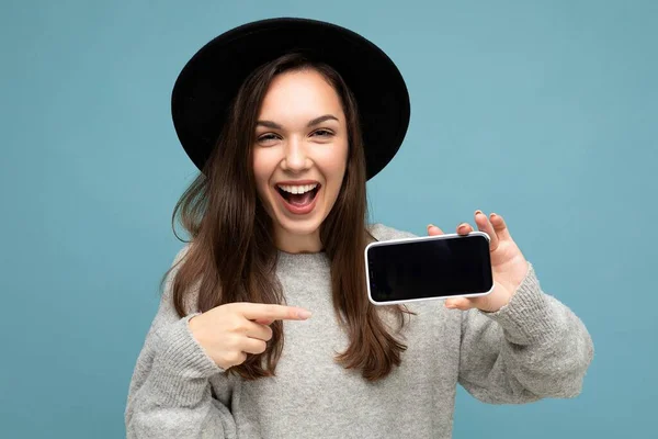 검은 모자와 회색 스웨터를 입고 있는 아름다운 젊은 여성의 초상화 사진 배경에서 분리 된 스마트폰을 보여 주는 스마트폰을 들고 있는 사진 — 스톡 사진