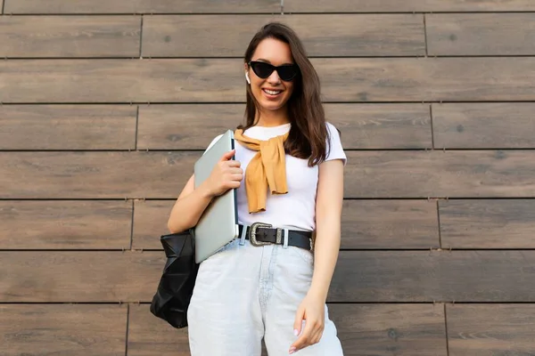 Beautiful smiling happy charming fascinating young brunet lady looking at camera holding computer laptop and black sunglasses in white t-shirt and light blue jeans in the street near brown wall — Stock Photo, Image