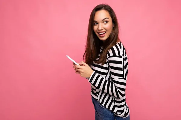 Side profile shot van mooie jonge glimlachende brunette vrouw met behulp van mobiele telefoon communiceren via sms dragen trui geïsoleerd op muur achtergrond kijken naar de zijkant — Stockfoto