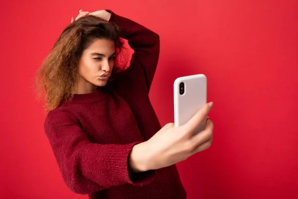 Beautiful young woman with curly hair wearing dark red sweater isolated on red background wall holding and using smart phone looking at telephone screen and taking selfie and giving kiss — Stock Photo, Image