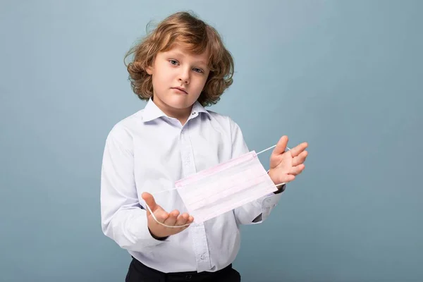 Guapo rubio rizado niño pequeño sosteniendo máscara protectora de pie aislado sobre fondo azul. Concepto covid — Foto de Stock