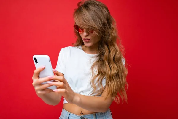 Photo of amazed beautiful young woman holding mobile phone wearing sunglasses everyday stylish outfit isolated over wall background looking at device screen and communicating — Stock Photo, Image