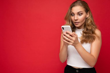 Şaşırdım, beyaz tişört giyen güzel sarışın kadın kırmızı arkaplan üzerinde izole edilmiş akıllı telefon ve cep telefonuyla mesajlaşarak gadjet ekranına bakıyor.