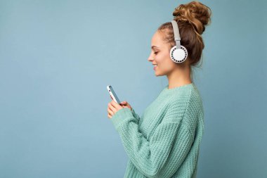 Güzel, neşeli, güler yüzlü, şık kıyafetler giyen, arka plan duvarından izole edilmiş, cep telefonu kullanan, beyaz bluetooth kulaklık takan ve müzik dinleyen genç bir kadının fotoğrafı.