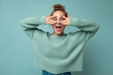 Genç, mutlu, neşeli, samimi duyguları olan, mavi renkli kazak giyen, fotokopi alanı ile izole edilmiş, gözleri ellerle kaplayan ve eğlenen güzel bir sarışın kadının fotoğrafı.