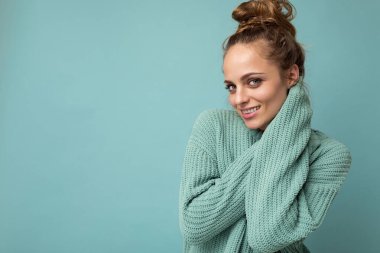 Genç, güzel, kıvırcık saçlı, seksi bir yüzü olan, neşeli ve mutlu yüzlü, modaya uygun mavi süveter giyen, mavi arka planda izole edilmiş, fotokopi alanı olan bir kadın.