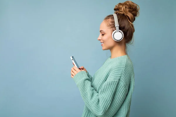 멋진 미소를 짓고 있는 아름다운 젊은 여성의 사진 이 배경 벽에 고립되어 있고, 음악을 듣고 있는 흰색 블루투스 헤드폰을 쓰고 있는 휴대 전화를 사용하고 있다. — 스톡 사진