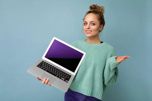 Frumoasă blondă întunecată tânără femeie cu păr creț adunat uitându-se la camera care deține laptop-ul computerului cu ecran de monitor gol cu batjocură în sus și spațiu de copiere purtând mânecă lungă albastră izolată pe albastru deschis — Fotografie, imagine de stoc