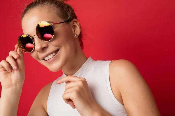Photo shot of beautiful smiling positive young blonde woman wearing casual clothes and stylish sunglasses isolated over colorful background looking at camera — Stock Photo, Image