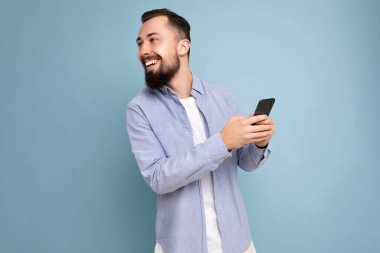 Gülümseyen, genç, esmer, sakallı, şık beyaz tişörtlü ve mavi gömlekli bir adamın portresi. Mavi arka planda, elinde boş bir alan var ve telefon kullanıyor.
