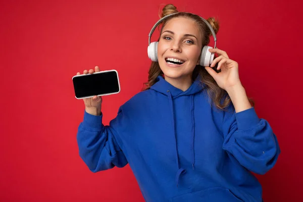 Hermosa mujer joven sonriente feliz con elegante atuendo casual aislado en la pared de fondo sosteniendo y mostrando el teléfono móvil con pantalla vacía para maqueta usando auriculares bluetooth blancos — Foto de Stock
