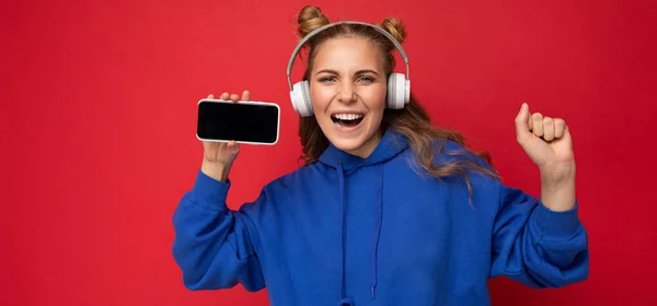 Foto panorâmica de fascinante feliz sorrindo jovem vestindo elegante capuz azul isolado sobre fundo vermelho vestindo fones de ouvido sem fio bluetooth branco e ouvir música e mostrando móvel — Fotografia de Stock