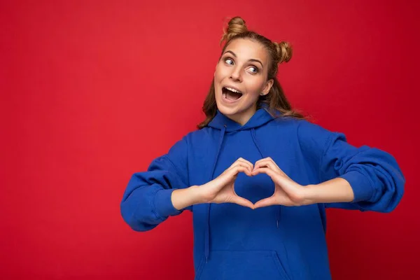 Νεαρή ευτυχισμένη θετική ελκυστική ξανθιά γυναίκα με δύο κέρατα με ειλικρινή συναισθήματα φορώντας κομψό φωτεινό μπλε hoodie απομονώνονται σε κόκκινο φόντο με χώρο αντίγραφο και δείχνει χειρονομία καρδιάς — Φωτογραφία Αρχείου