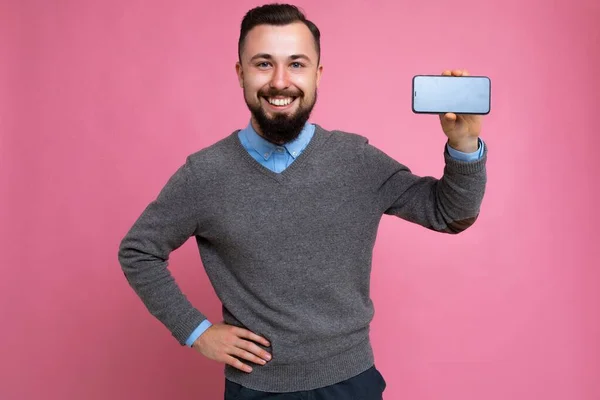 Sorrindo feliz bonito feliz legal jovem morena homem sem barba com barba vestindo elegante camisola cinza e camisa azul de pé isolado sobre rosa parede de fundo segurando smartphone e mostrando telefone — Fotografia de Stock