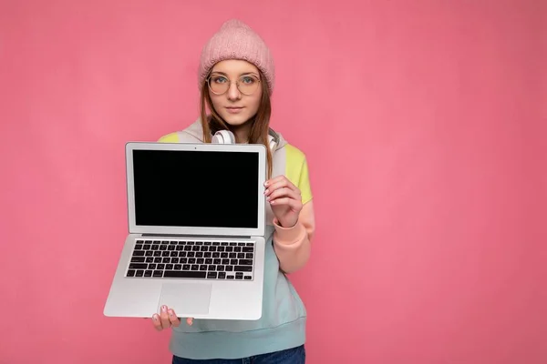예쁜 젊은 여성 이 모자 색깔의 스웨터를 입고 안경을 끼고 빈 복사기가 달린 컴퓨터 노트북을 들고 흰색 헤드폰을 끼고 핑크 벽 배경에 고립된 카메라를 보고 있습니다 — 스톡 사진
