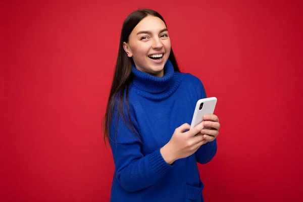 Aantrekkelijk positief uitziende jonge brunette vrouw dragen stijlvolle blauwe warme trui evenwicht geïsoleerd op rode achtergrond met lege ruimte in de hand en met behulp van mobiele telefoon messaging sms op zoek — Stockfoto