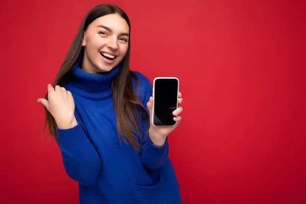 Портрет улыбающейся красивой счастливой молодой брюнетки, одетой в случайный синий свитер, изолированный на красном фоне с пустым пространством, держа в руке мобильный телефон и показывая смартфон с пустым экраном — стоковое фото