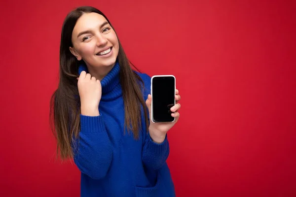 Closeup of delightful beautiful happy young brunette woman wearing casual blue sweater isolated over red background with empty space holding in hand mobile phone and showing smartphone with empty — Stock Photo, Image