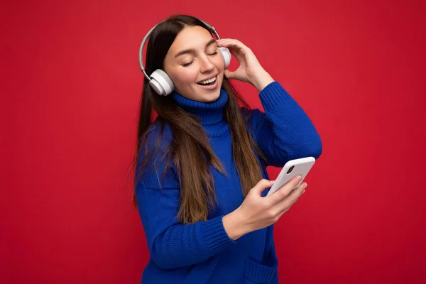 Zdjęcie pięknej, radosnej, uśmiechniętej młodej kobiety w stylowych, luźnych ciuchach odizolowanych nad ścianą tła, trzymającej i używającej telefonu komórkowego w białych słuchawkach bluetooth słuchających muzyki i — Zdjęcie stockowe