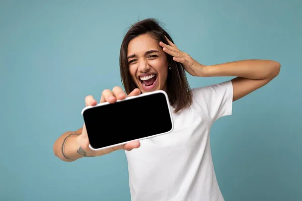 흰색 티셔츠를 입고 푸른 배경에 고립된 모습을 한 젊은 브라 운트 여성이 빈 화면으로 스마트폰을 보여 주는 휴대폰을 들고 핸드폰을 들고 있는 사진. — 스톡 사진