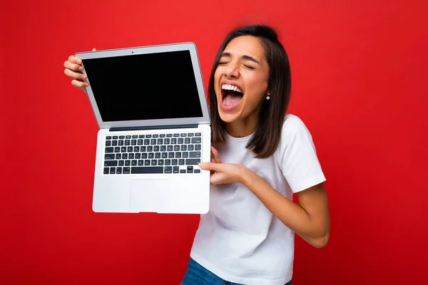 Portret de aproape de frumos fericit tanara bucuros cu tunsoare bruneta scurta tinand laptopul computerului cu ochii inchisi purtand tricou alb si blugi izolati pe fundalul peretelui rosu — Fotografie, imagine de stoc