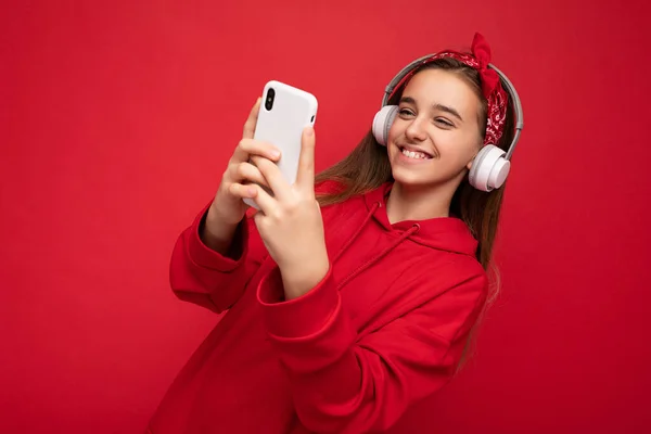 긍정적 이고 매력적 인 갈색 머리 여자붉은 후드를 착용하고 빨간 배경에서 고립되어 있고 스마트폰을 사용하여 온라인으로 대화하고 있다. — 스톡 사진