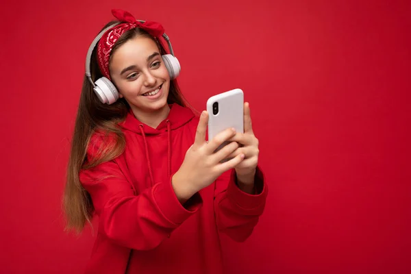 긍정적 인 미소를 가진 빨간 후드를 입고 빨간 배경에서 고립된 갈색 머리 소녀가 스마트폰으로 온라인에서 흰색 무선 헤드폰을 쓰고 음악을 듣고 있다. — 스톡 사진