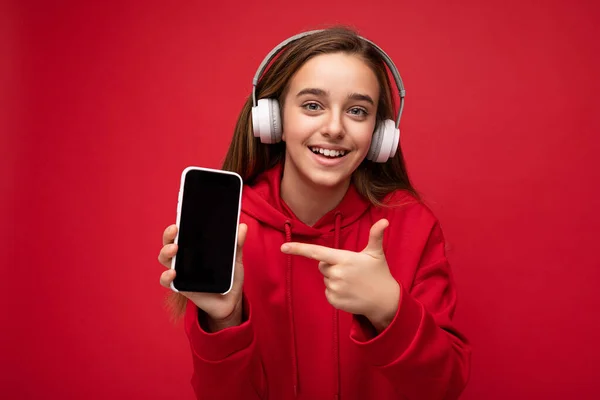 Uśmiechnięta piękna brunetka nastolatka kobieta w czerwonej bluzie izolowane na czerwonym tle gospodarstwa i pokazując smartfon z pustym wyświetlaczem do cięcia noszenia białych słuchawek bezprzewodowych słuchania — Zdjęcie stockowe