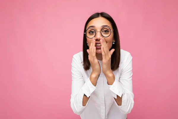 Porträtt av unga positiva glada attraktiv brunett kvinna med uppriktiga känslor bär casual vit skjorta och optiska glasögon isolerade över rosa bakgrund med kopia utrymme och säger hemlighet — Stockfoto