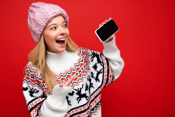 Photo d'une jolie jeune femme blonde souriante portant un chapeau tricoté chaud et un pull chaud d'hiver isolé sur fond rouge montrant un smartphone avec écran vide regardant l'affichage de l'appareil — Photo