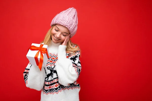 Foto skott av vackra glada leende förvånad ung blond kvinna isolerad över röd bakgrund vägg bär vinter tröja och rosa hatt håller vit presentförpackning med rött band — Stockfoto