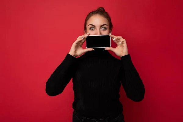 Foto de atraente positivo jovem morena vestindo camisola preta de pé isolado sobre fundo vermelho mostrando telefone celular com tela vazia para mockup olhando para a câmera — Fotografia de Stock