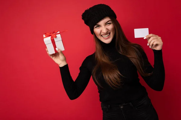 Vacker glad rolig arg ung brunett kvinna bär svart tröja och hatt isolerad på röd bakgrund innehar kreditkort och presentförpackning tittar på kameran — Stockfoto