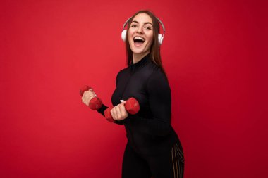 Çekici, gülümseyen, esmer, siyah spor giysiler giyen, kırmızı arka plan duvarında izole edilmiş, halterli, beyaz kulaklık takan, iyi müzik dinleyen, esmer bir kadın.