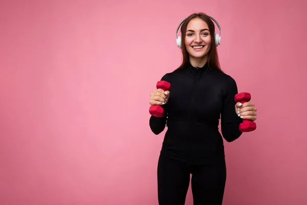 Pretty positive smiling young brunette woman wearing black sport clothes isolated over pink background wall doing fitness using dumbbells wearing white bluetooth headphones listening to good music and — Stock Photo, Image