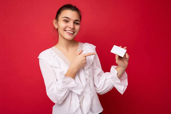 Фото красивой позитивной улыбающейся молодой темнокожей блондинки в белой блузке на красном фоне, держащей кредитную карту, смотрящей в камеру и указывающей пальцем на пластиковую бесконтактную карту — стоковое фото