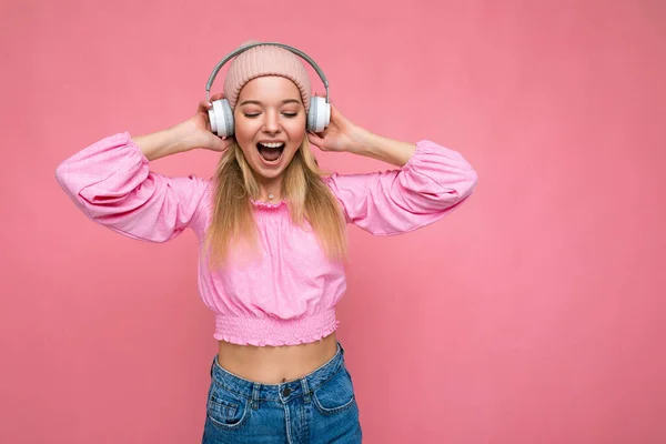 Attractive positive emotional smiling young blonde woman wearing pink blouse and pink hat isolated over pink background wall wearing white wireless bluetooth earphones listening to music having fun