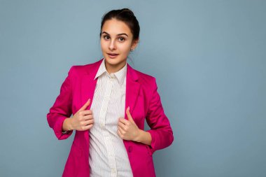 Çekici, çekici, kendine güvenen, samimi duyguları olan beyaz gömlekli ve mavi arka planda izole edilmiş pembe ceketli genç, güzel, güzel esmer kız öğrencinin fotoğrafı.