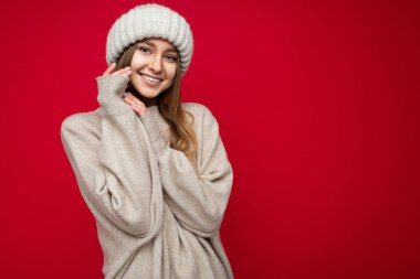 Çekici, mutlu, gülümseyen, koyu saçlı, kırmızı arka plan duvarında izole bir şekilde duran, bej süveter ve kış bej şapkalı, kameraya bakan bir kadın.