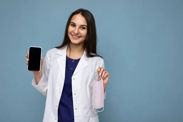 Photo de belle jeune femme brune souriante belle vêtue d'un manteau médical blanc isolé sur fond bleu avec espace de copie tenant smartphone montrant le téléphone à la main avec écran vide — Photo