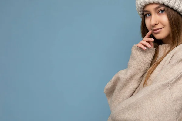 Closeup shot of attractive happy sexy young dark blonde woman standing isolated on blue background wall wearing beige warm sweater and winter beige hat looking at camera and dreaming — Stock Photo, Image