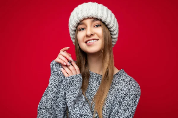Portrét fotografie krásné sexy usmívající se mladé tmavé blondýny žena stojící izolované přes červené pozadí zdi na sobě šedý svetr a zimní béžový klobouk při pohledu na kameru — Stock fotografie