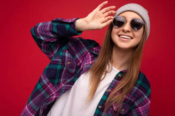 Detailní fotografie okouzlující mladé blondýny izolované přes červené pozadí na sobě barevné stylové košile šedý klobouk a moderní sluneční brýle při pohledu do kamery — Stock fotografie