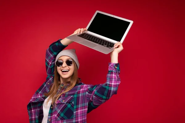 Uimitor fermecător frumos râzând amuzant zâmbind blondă tânără care deține laptop-ul computerului cu ecran de monitor gol purtând pălărie de ochelari de soare și cămașă colorată uitându-se la camera izolată peste roșu — Fotografie, imagine de stoc