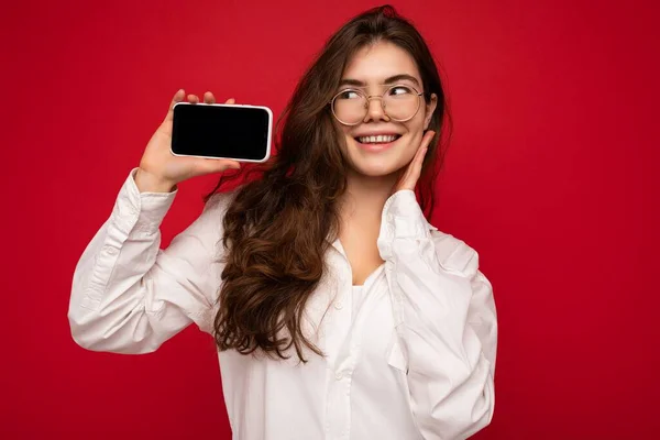 흰색 셔츠와 광학 안경을 단 매력적 인 젊은 브루넷 여성 이 손에 들고 빨간 배경 위에 고립된 채웃으며 사진을 찍기 위해 빈 화면을 가진 휴대 전화를 보여 주고 있다. — 스톡 사진