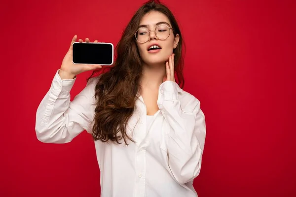 Foto van sexy mooie jonge brunette vrouw dragen witte shirt en optische bril geïsoleerd over rode achtergrond houden in de hand en toont mobiele telefoon met leeg scherm voor cutout kijken naar — Stockfoto