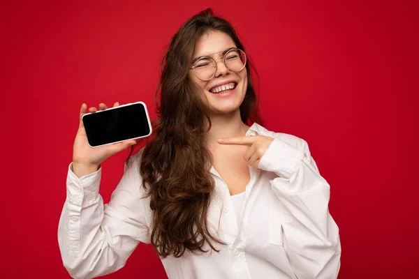 Sorrindo feliz atraente jovem morena vestindo camisa branca e óculos ópticos isolados sobre fundo vermelho segurando na mão e mostrando telefone celular com tela vazia para maquete — Fotografia de Stock