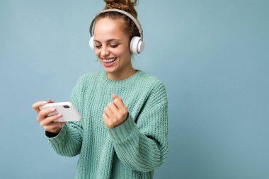 Mavi arka plan üzerinde izole edilmiş mavi süveter giyen, beyaz bluetooth kulaklık takan, müzik dinleyen ve cep telefonu kullanan çekici, duygusal pozitifliğin yan plan fotoğrafı.